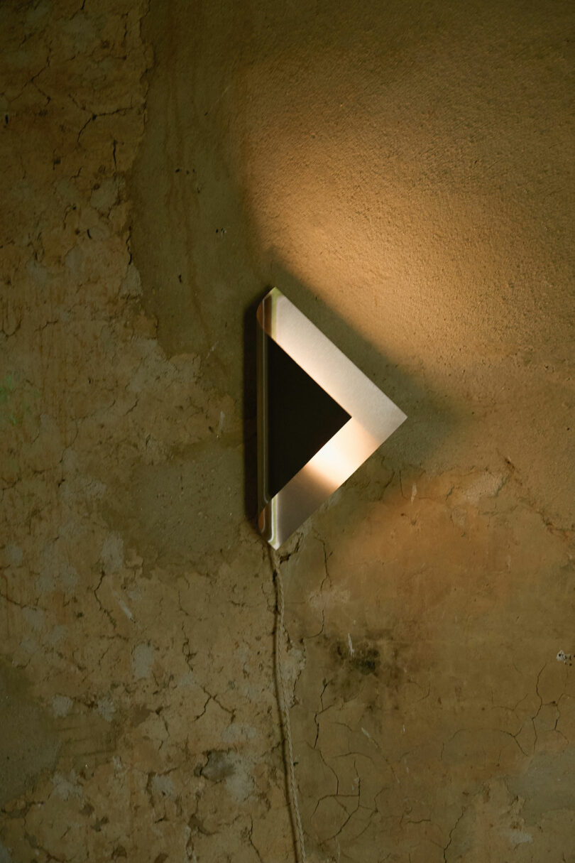 A triangular wall light fixture is mounted on a cracked, textured wall, emitting soft light upwards and outwards.