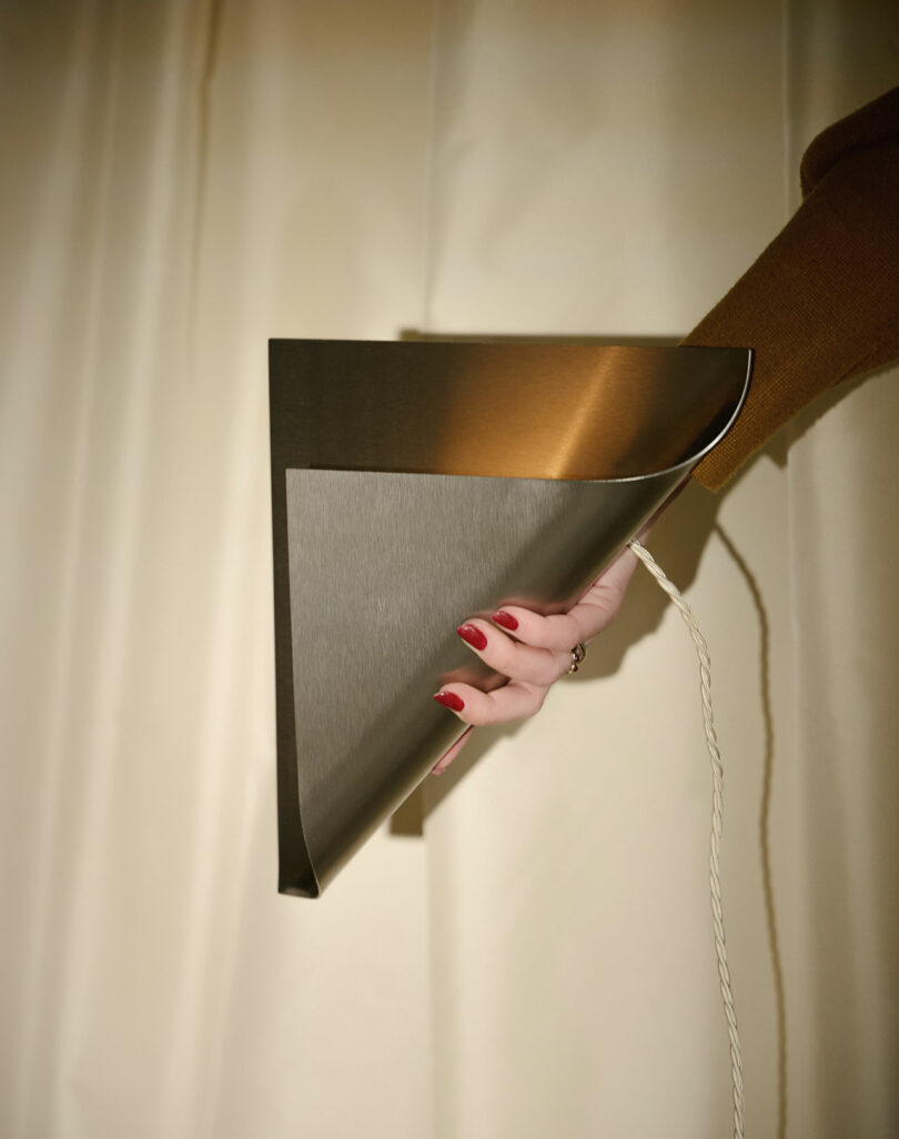 A hand with red nails holds a metallic, folded sheet-like object with a white cord against a neutral curtain background.