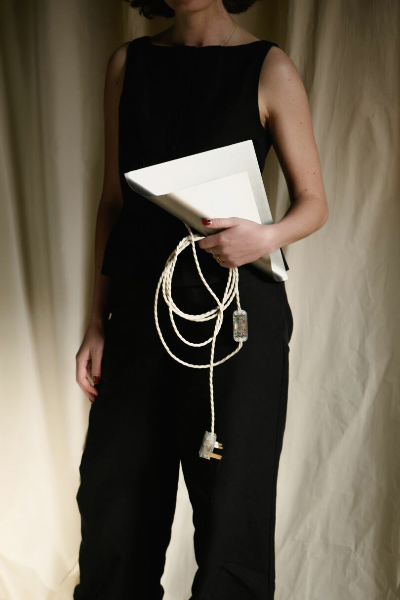 A person in black clothing holds a closed laptop and a coiled power cable in one hand, standing against a neutral curtain backdrop.