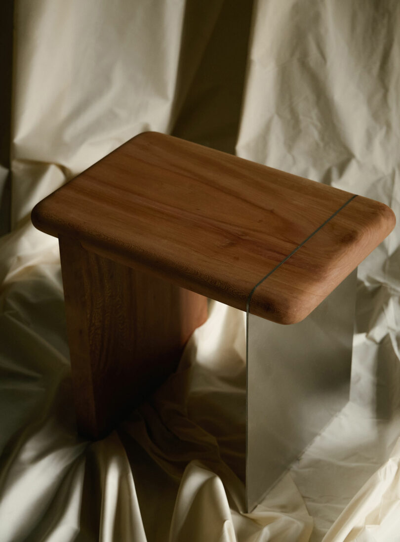 A small wooden side table with a rectangular top, a wooden leg on one side, and a transparent acrylic leg on the other, placed on draped cream-colored fabric.