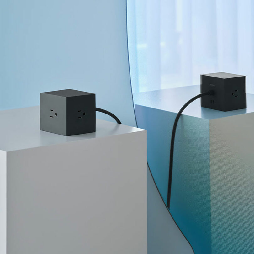 A black, cube-shaped power outlet sits on a white pedestal, showcasing sleek technology, with its reflection visible in a nearby mirror.