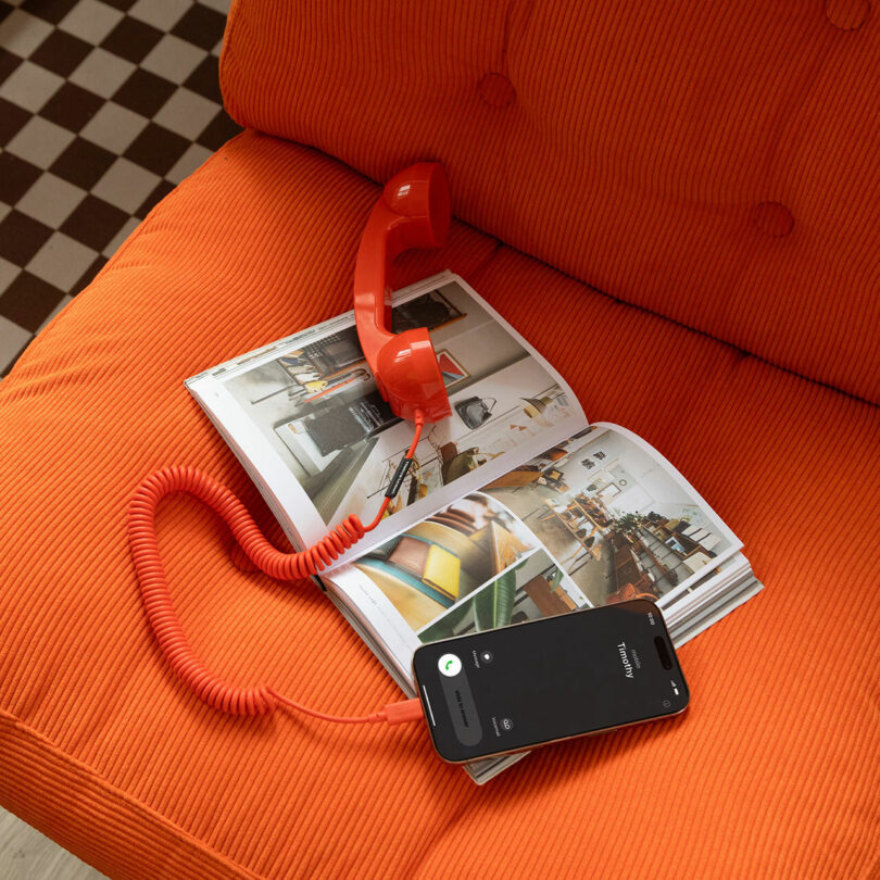 A red corded telephone handset and a smartphone rest on an open magazine on an orange couch; the smartphone’s incoming call screen highlights the blend of classic and modern technology.