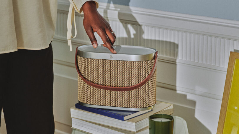 A person touches the top of a woven portable speaker with a leather handle—a stylish piece of technology—placed on stacked books next to a green candle and a yellow-framed picture.