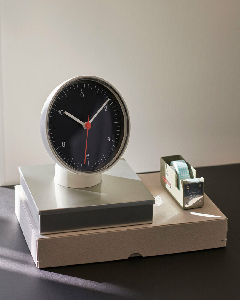 A round analog clock showing 8:02 sits on stacked boxes next to a small box with a tape dispenser, making a stylish addition for any Gift Guide 2025 display on a dark surface.