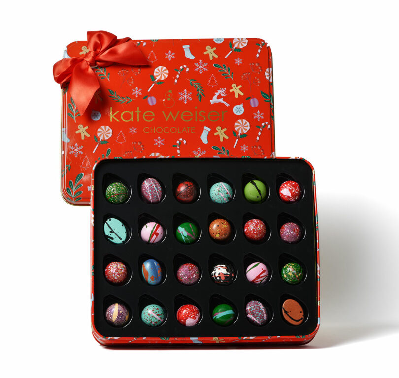 A festive red tin box with a bow holds 24 assorted, decorated chocolate bonbons arranged in four rows. Featured in the Gift Guide 2025, its lid displays holiday-themed illustrations and the text "kate weiser chocolate.