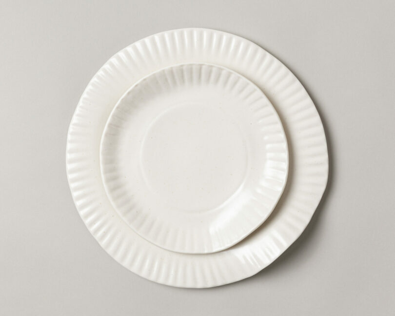 Two white ceramic plates with a fluted edge are stacked on a neutral gray background.