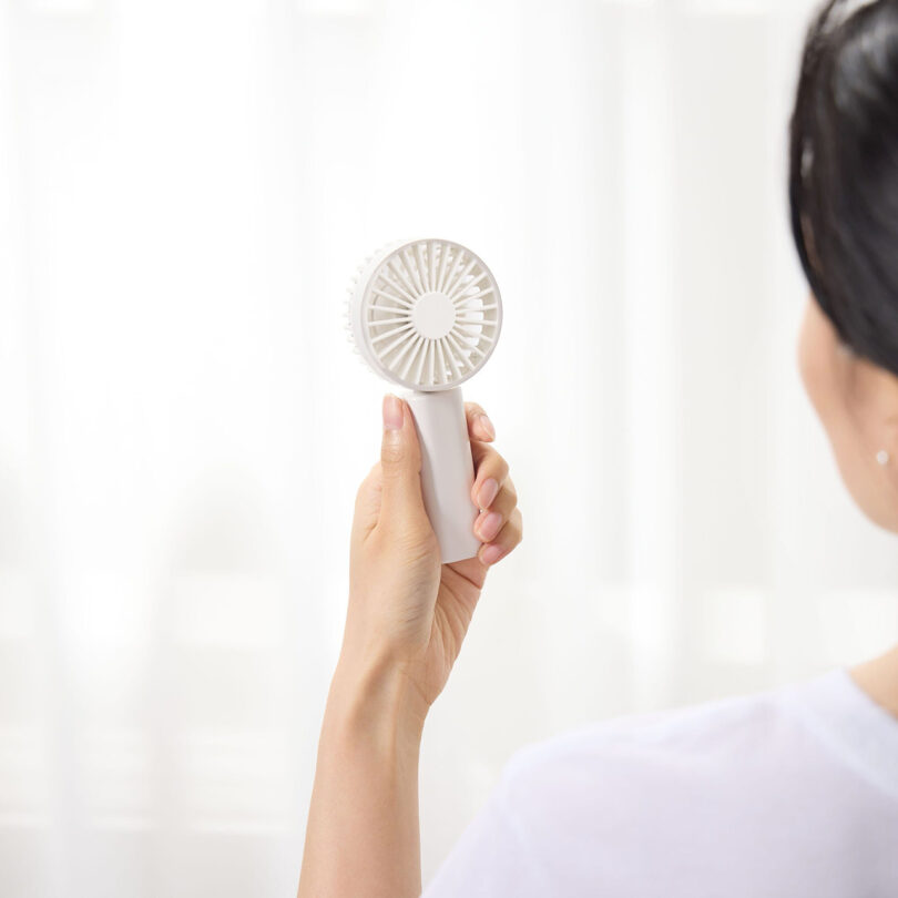 A person is holding a small, white handheld fan in front of a sheer curtain in a bright room—an ideal pick for the 2025 Gift Guide.