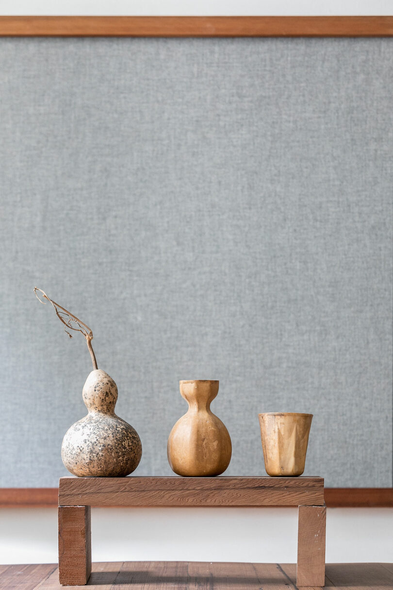 Three wooden vessels, one holding a dried branch, are displayed on a small wooden platform in front of a gray fabric-covered board.