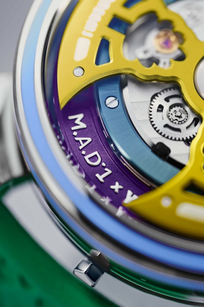 Close-up of a wristwatch with visible mechanical gears, yellow and purple components, and the engraving "M.A.D.1 x" on the purple part of the watch face