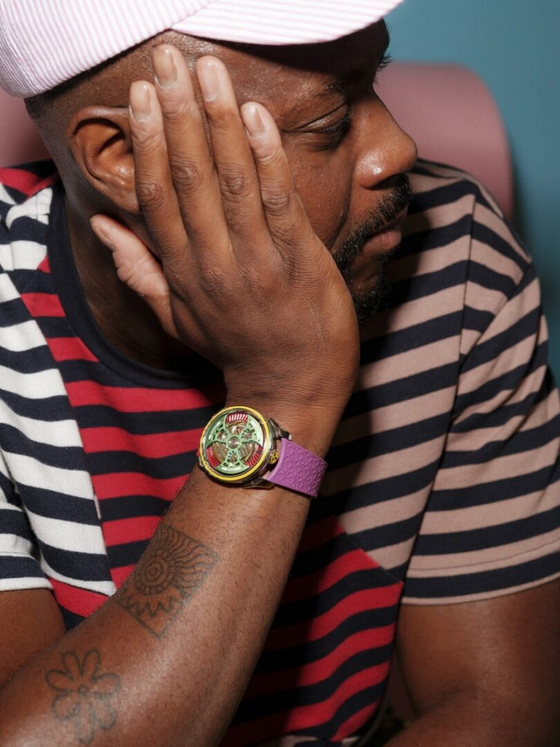 A person wearing a striped shirt and cap rests their face on their hand, displaying a colorful wristwatch and a geometric tattoo on their forearm