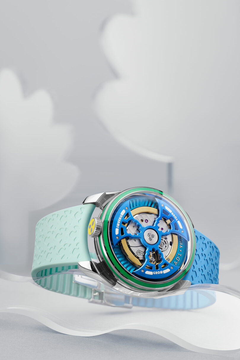 A wristwatch with a blue and light green strap, featuring a visible mechanical movement and a circular, open-faced blue dial, is displayed against a soft, abstract background