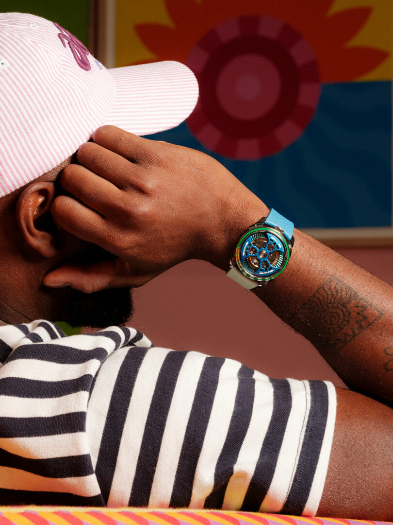 A person wearing a striped shirt and pink cap rests their arm on a surface, displaying a blue wristwatch; colorful artwork is in the background