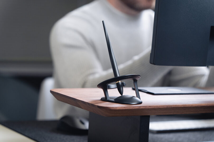 A Novium Hoverpen in its stand sits on a wooden desk next to a monitor, with a person in a light sweater working in the background.