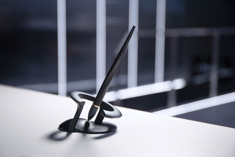 A modern black Novium Hoverpen stands upright in a sculptural black holder on a white surface, with blurred lights and a dark background behind.