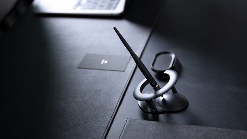 A Novium Hoverpen digital stylus rests vertically in a circular holder on a black desk, with a laptop and a card visible in the background.