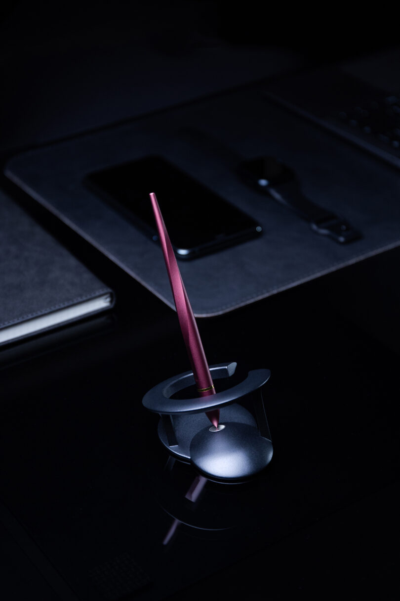 A metallic red Novium Hoverpen stylus is placed upright in a modern holder on a dark desk, with a smartphone, smartwatch, and closed laptop in the background.