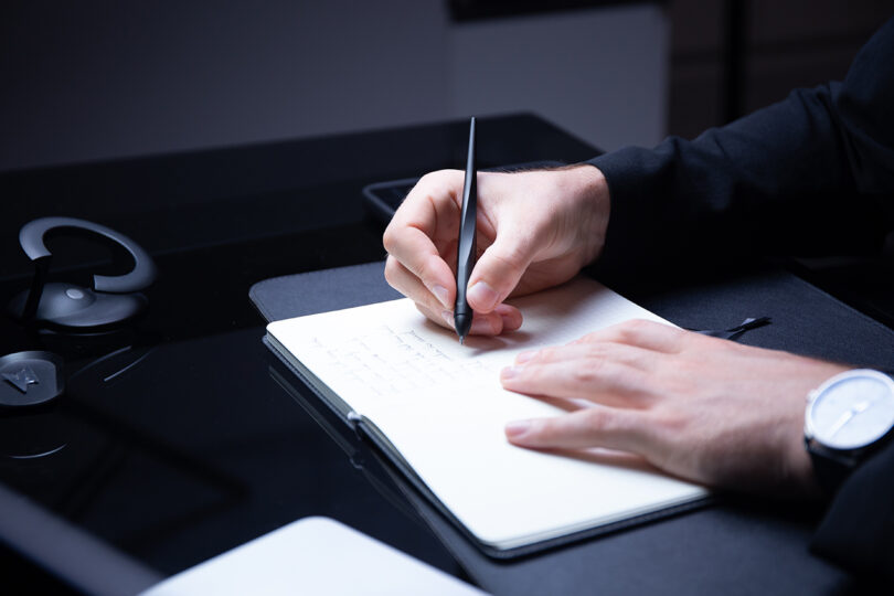 A person writes in a notebook with a Novium Hoverpen at a black desk, wearing a watch and a dark long-sleeve shirt.