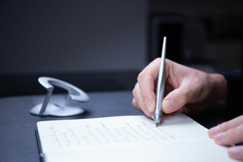 A person writes with a silver pen in a notebook on a desk, while the sleek Novium Hoverpen, a modern circular object, elegantly floats in the background.
