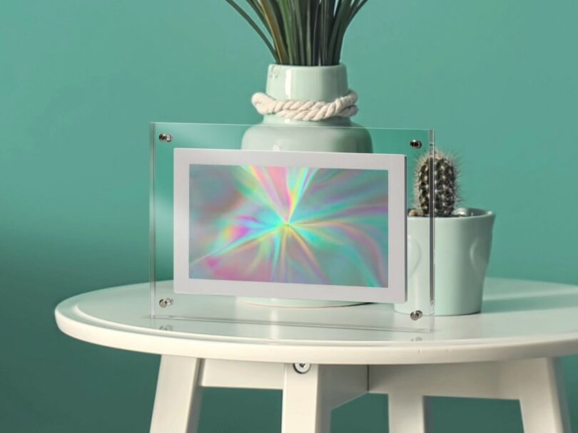A holographic print in a clear acrylic frame sits on a white round table, with a potted plant and a small cactus in the background against a teal wall