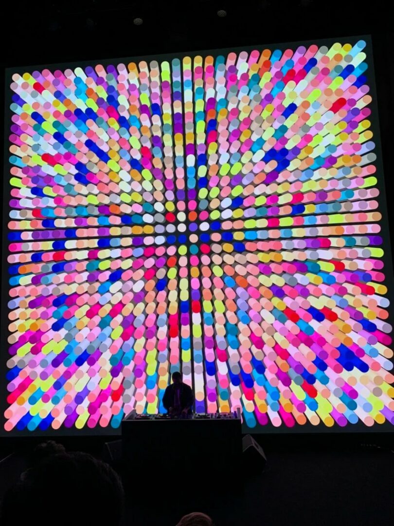 A person stands at a DJ booth in front of a large, brightly colored digital display featuring a radiating pattern of circular shapes