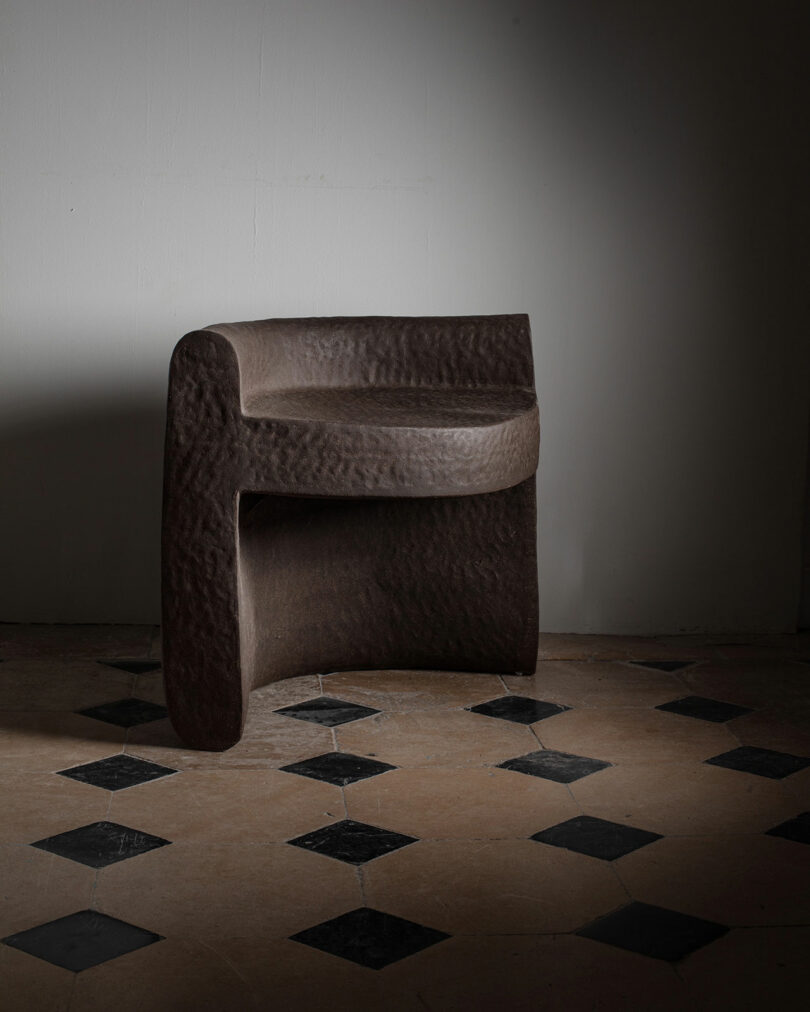 A sculpted, dark brown chair by Kelsie Rudolph with a textured surface sits on a checkered tile floor against a plain white wall.