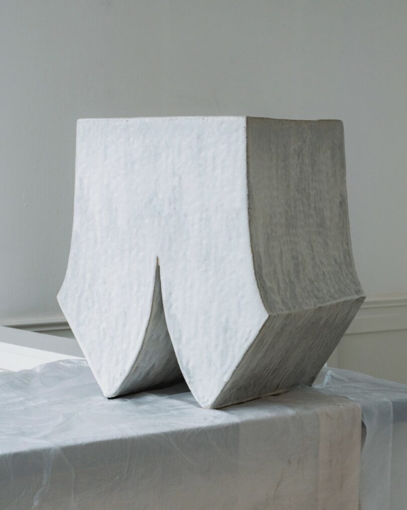 A geometric, white sculptural object with two downward-pointing curves sits on a draped table against a plain background