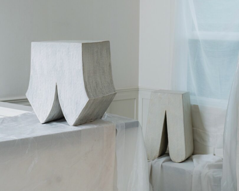 Two geometric, gray sculptural objects sit in a minimalist room draped with translucent white fabric. One object is on a table, the other is on the floor near a window