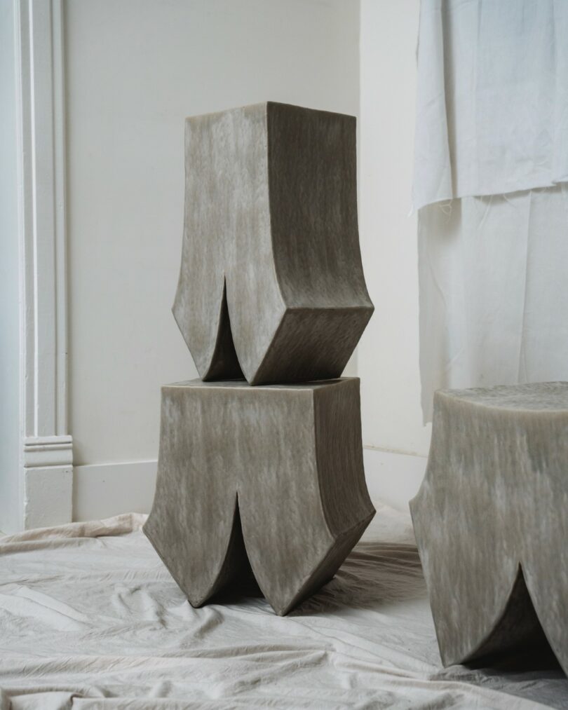 Two sculptural, gray, geometric stools with flared bases are stacked vertically on a draped fabric floor in a minimalist room with white walls and a hanging sheet
