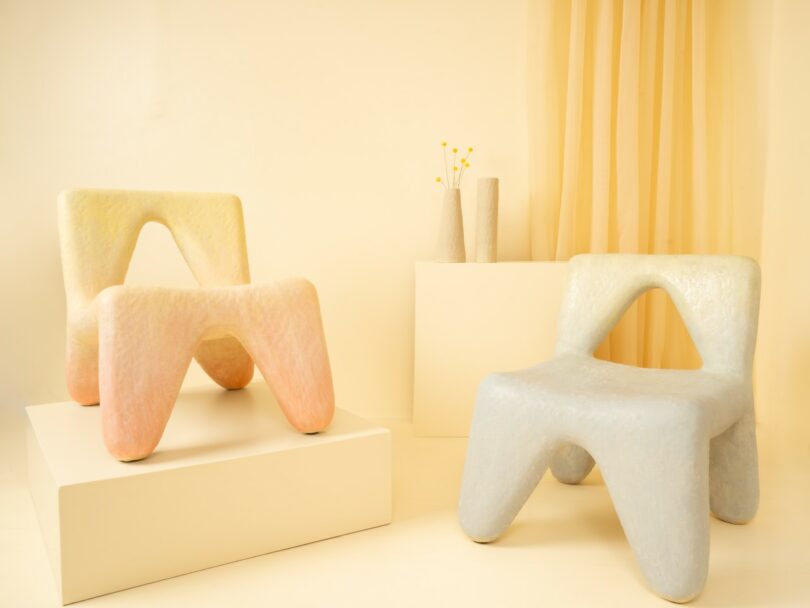 Two modern, sculptural chairs in pastel colors are displayed in a minimal setting with a white plinth, neutral background, and two vases holding yellow billy ball flowers