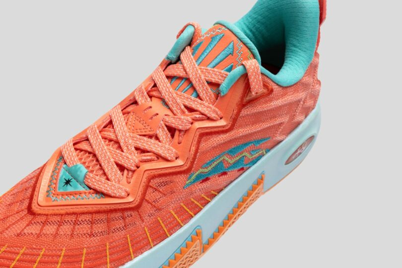 Close-up of an orange athletic shoe with blue and yellow accents, showing detailed stitching and textured sole against a plain gray background