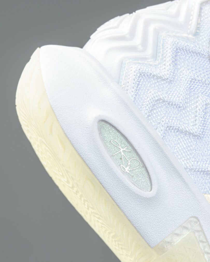 Close-up of a white athletic sneaker with textured and glossy detailing, showing the laces, tongue, and part of the upper design against a plain gray background.