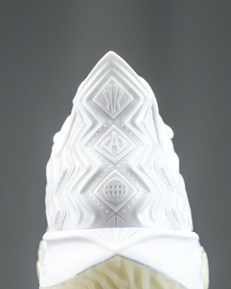 Close-up of a white athletic sneaker with textured and glossy detailing, showing the laces, tongue, and part of the upper design against a plain gray background.