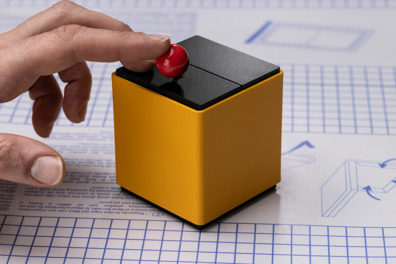 A hand presses a small red button on top of a yellow and black cube, placed on graph paper with technical drawings and instructions.