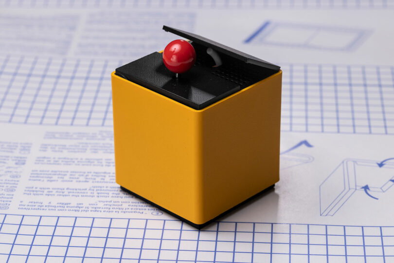 A small yellow cube with a black top, featuring a hinged lid partly open to reveal a red joystick, placed on graph paper with printed text and diagrams.