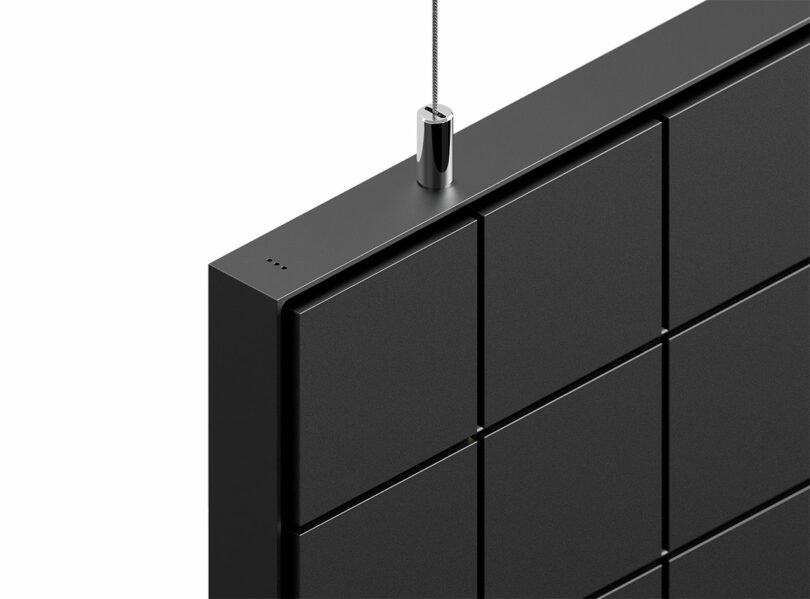 Close-up of the top corner of a black, grid-patterned panel suspended by a thin cable attached to a metal fitting.