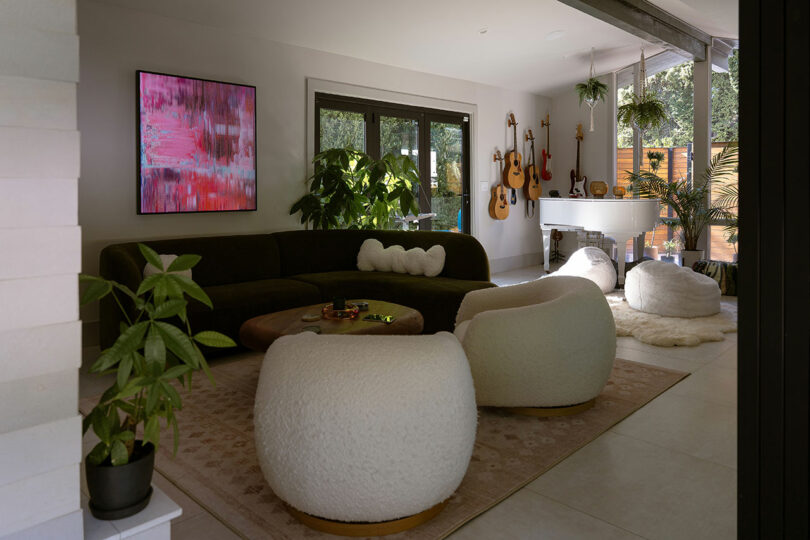 Modern living room with a curved sofa, round white chairs, indoor plants, wall-mounted guitars, abstract wall art, large rug, and a white grand piano near large windows.