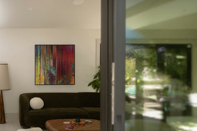 A modern living room with a dark green sofa, a round pillow, a standing lamp, a wooden coffee table, and a colorful abstract painting on the wall.