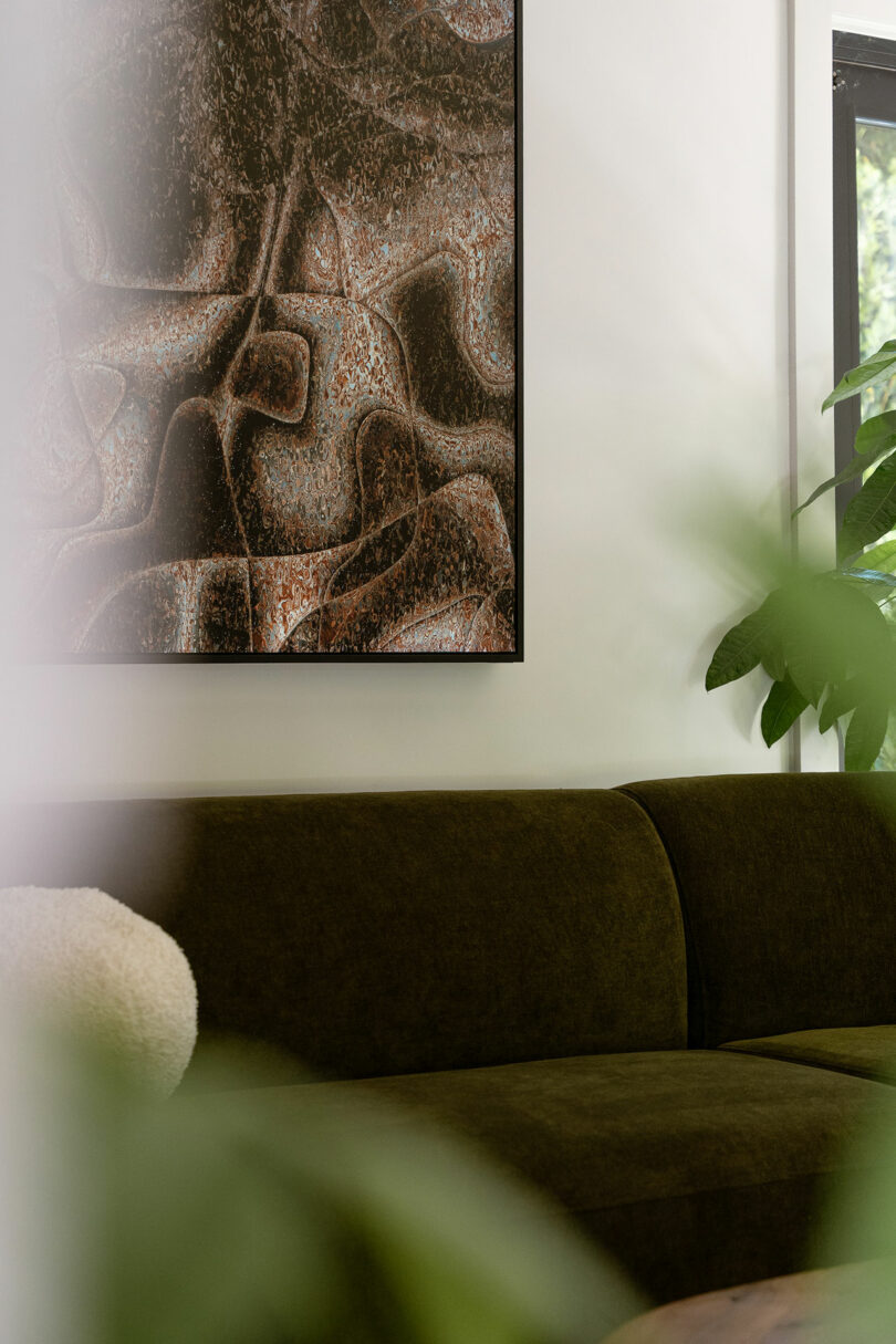 Green velvet sofa next to green plants, with an abstract artwork in earth tones hanging on the white wall above. Natural light enters from a window on the right.