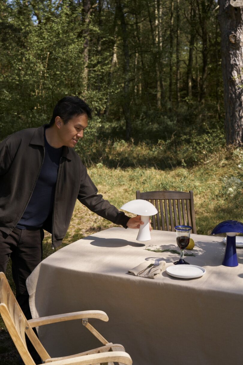 A man places a white lamp on an outdoor dining table set with plates, cutlery, glasses, and another blue lamp, surrounded by trees
