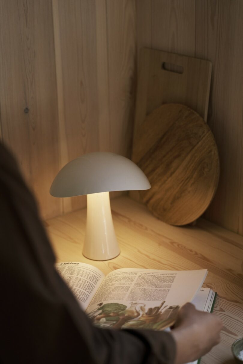A person reads a magazine at a wooden desk with a white mushroom-shaped lamp and two wooden cutting boards in the background