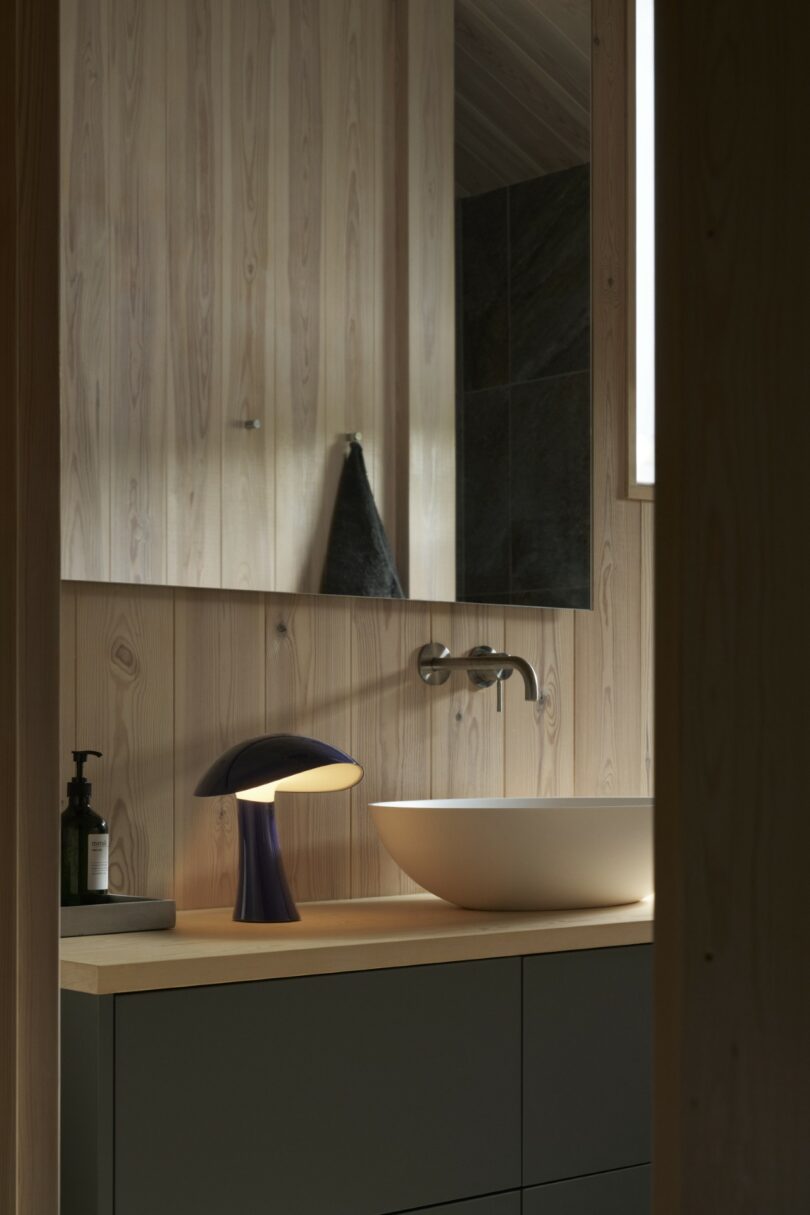 Modern bathroom with light wood walls, a round vessel sink, wall-mounted faucet, dark vanity, a black towel, a soap dispenser, and a purple table lamp on the counter