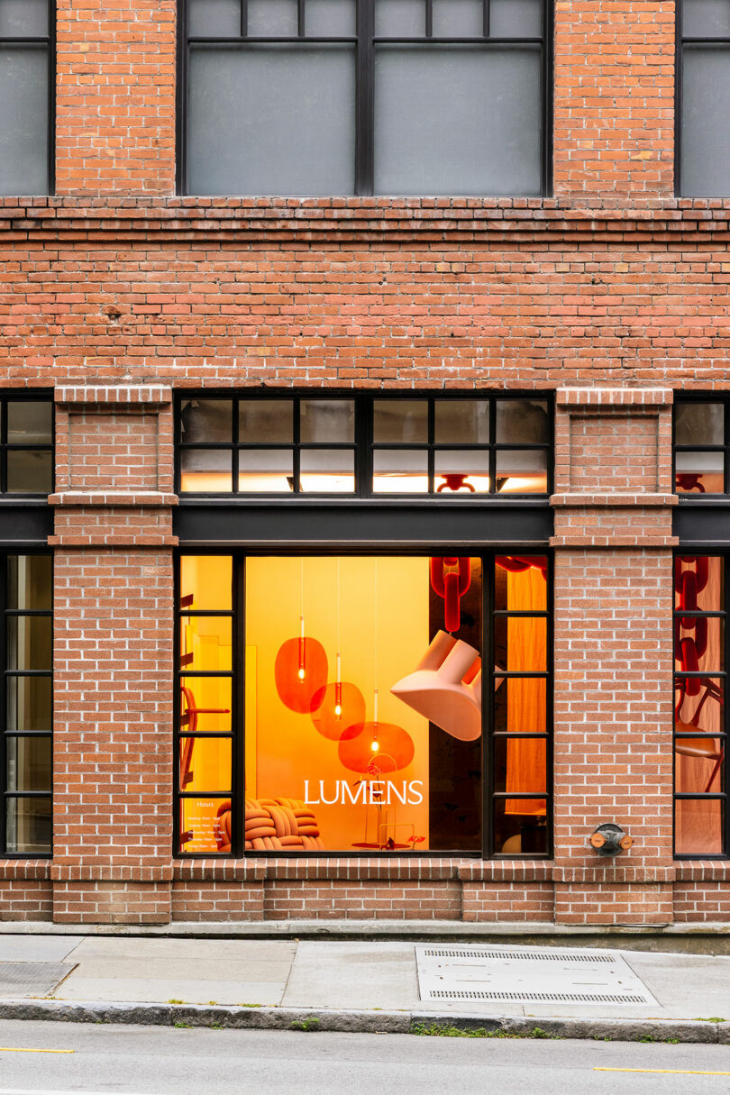 A brick building facade with a large window displaying modern orange pendant lights and the word "LUMENS" on the glass.