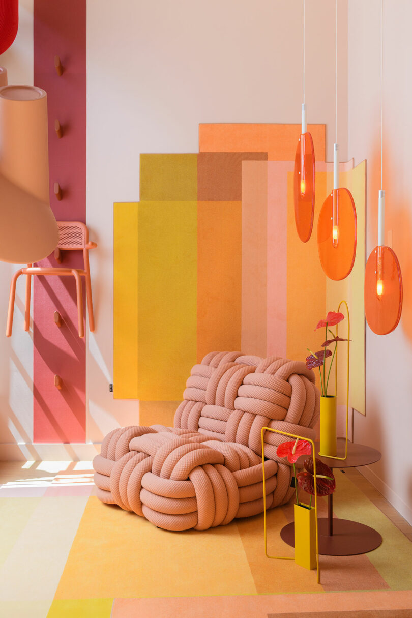 A modern room with geometric wall art, a textured pink lounge chair, a small table with vases, and three orange pendant lights. The décor features pink, yellow, and orange tones.