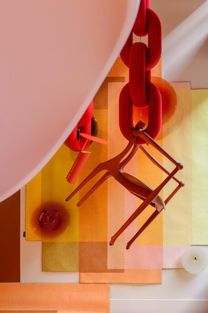 Overhead view of two red chairs suspended by large red links over layered rectangular rugs in orange, yellow, and pink tones, with glass vases on the floor.