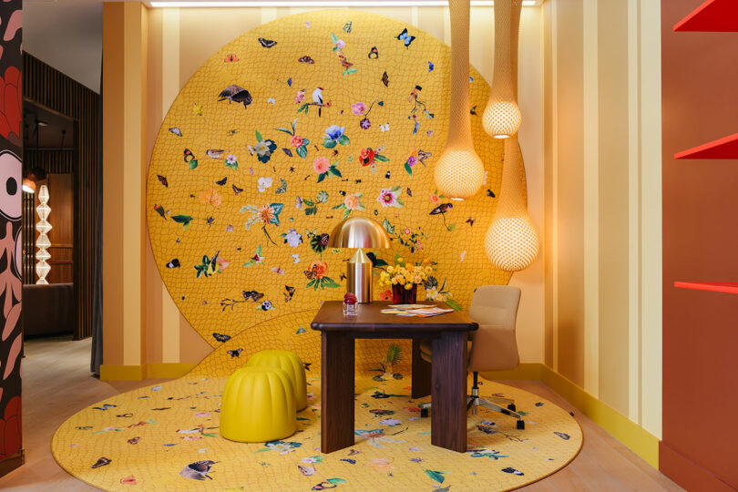 A colorful room with a yellow patterned wall and floor featuring flowers and butterflies, a wooden desk, yellow stools, a beige chair, and three pendant lights.