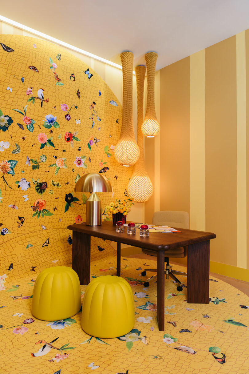 Modern office space with a wooden desk, gold lamp, yellow stools, and patterned yellow walls and floor with floral and butterfly designs, accented by hanging woven light fixtures.