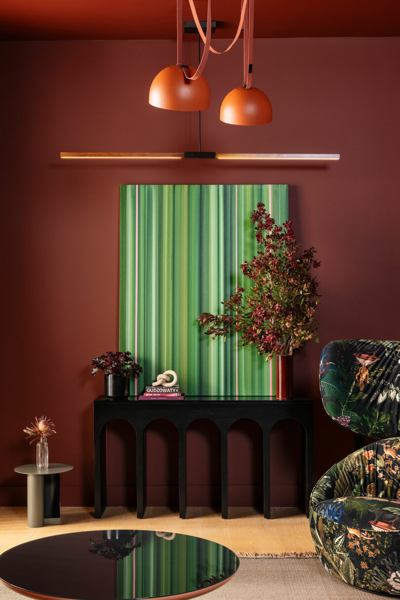 A modern living room with reddish-brown walls, orange pendant lights, green striped artwork, a black console table, and floral decor. A patterned armchair and tables complete the scene.