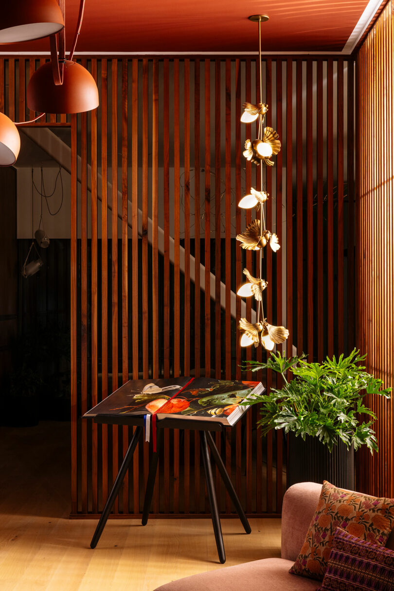 A modern interior with vertical wooden slats, a table displaying open books or artwork, a gold pendant light, a green potted plant, and a sofa with a patterned cushion.