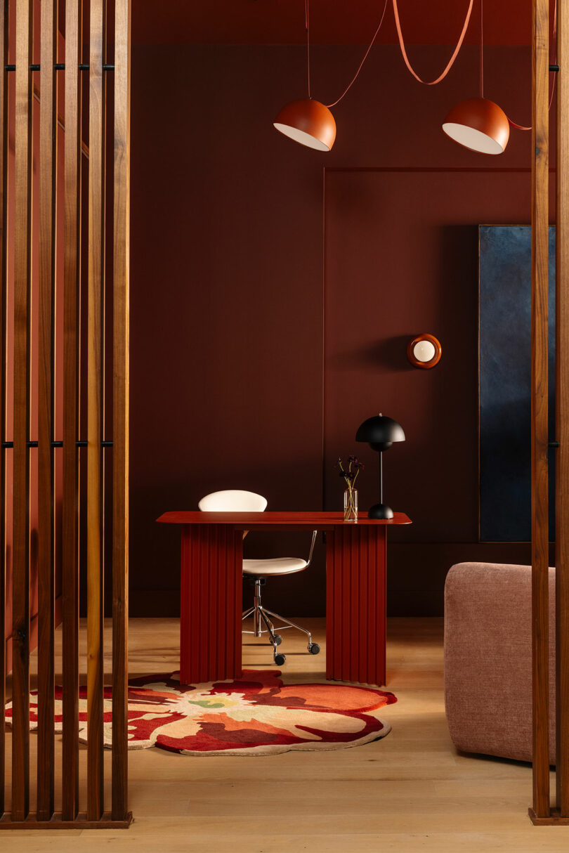 A minimalist office space features a red desk, white chair, black lamp, wall art, and geometric rug, with wooden slats and pendant lights under warm, muted red lighting.