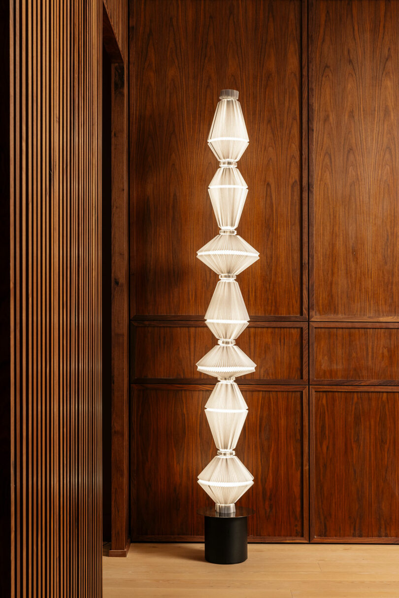 Tall sculptural floor lamp with stacked geometric white shades on a black base, positioned against wood-paneled walls in a modern interior.